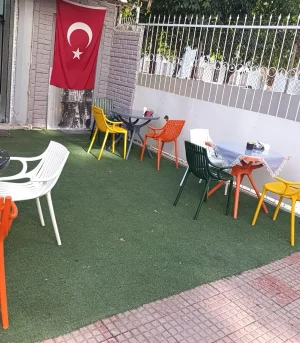 Kafe önündeki yuvarlak masaların çevresinde beyaz, yeşil, sarı ve turuncu plastik sandalye modelleri