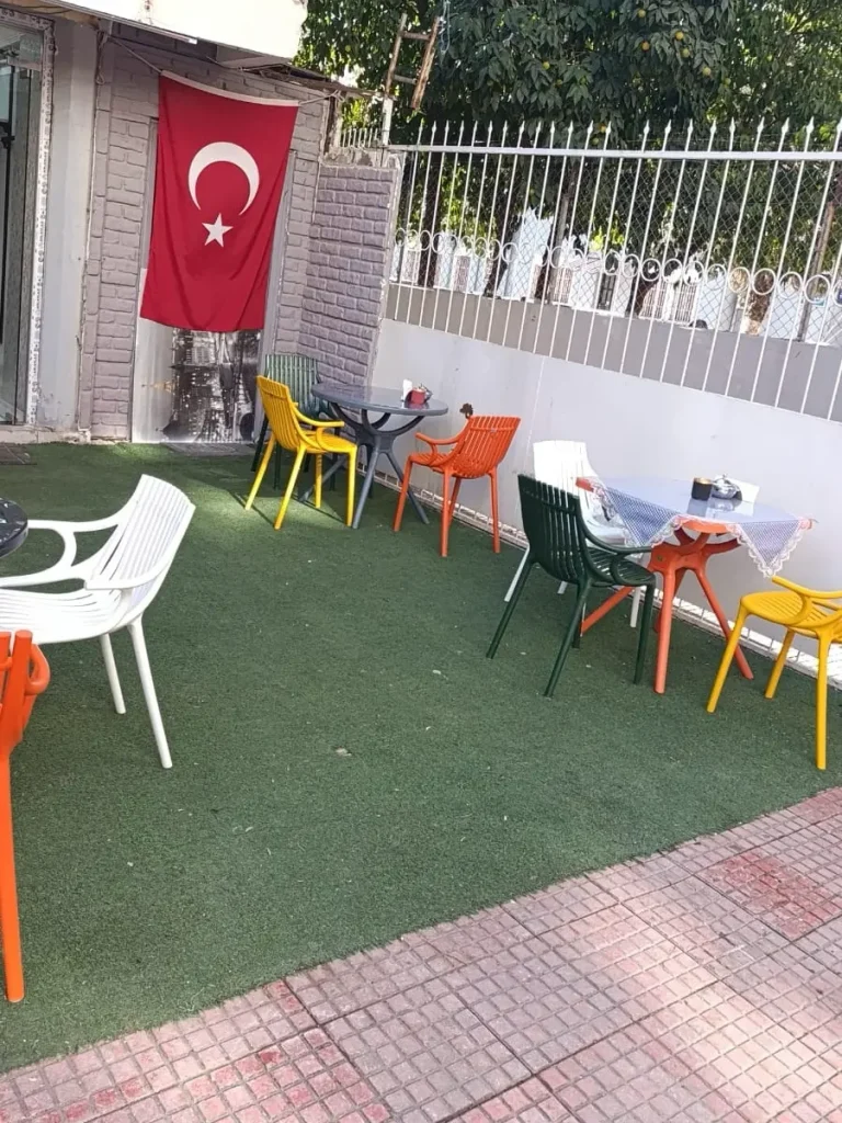 Kafe önündeki yuvarlak masaların çevresinde beyaz, yeşil, sarı ve turuncu plastik sandalye modelleri