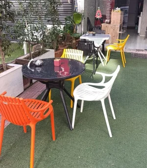 Yuvarlak siyah masa çevresinde turuncu ve beyaz kolçaklı, sarı kolçaksız plastik sandalyeler
